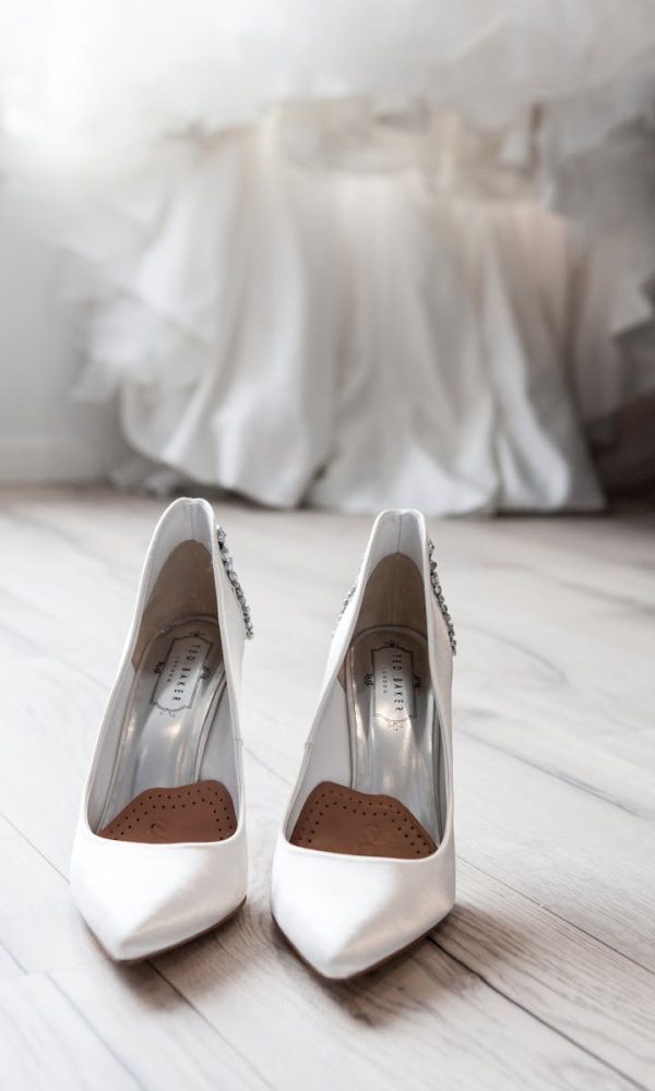 Stylish white bridal shoes positioned in front of a flowing wedding dress indoors.